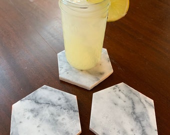 May include: Three white marble hexagon coasters with gray veining. A glass of lemonade with a lime wedge and a metal straw sits on one of the coasters.