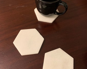 May include: Three white hexagonal coasters made of marble. Two coasters are laying flat on a dark brown wooden surface. A black coffee mug is sitting on the third coaster.