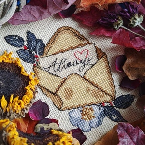May include: A cross-stitch embroidery of an envelope with the word "Always" written on it. The envelope is surrounded by flowers and leaves, and there is a red heart in the upper right corner.