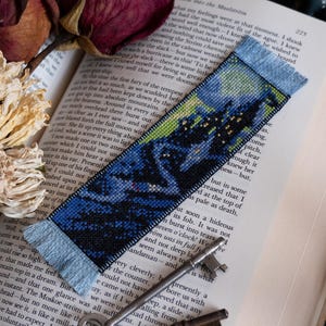 May include: A handmade bookmark with a cross-stitch design depicting a dark landscape with a starry sky. The bookmark has a blue fringe and is placed on an open book with dried roses and antique keys.