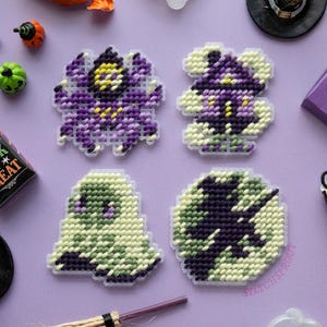 May include: Four plastic canvas needlepoint ornaments featuring Halloween designs: a purple spider, a purple witch hat, a light green ghost, and a black cat silhouette against a green moon. The pieces are stitched with yarn on clear plastic grids.