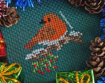 May include: A cross-stitch design of a robin perched on a branch with red berries, set against a green background. The image is surrounded by Christmas decorations, including gift boxes, pine cones, and blue tinsel. The text "@stitchsprout" is visible.