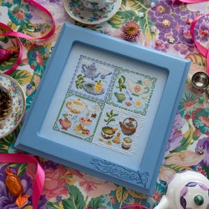 May include: A framed cross-stitch sampler featuring four tea-themed designs in a light blue frame. The sampler includes images of teapots, teacups, and tea-related items. The frame is surrounded by floral fabric, pink ribbons, and a teacup.