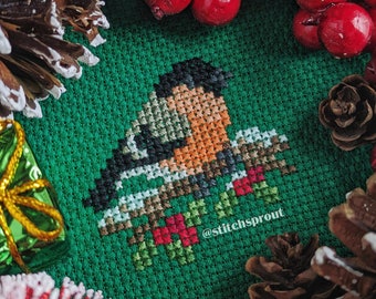 May include: Cross-stitch pattern of a red-breasted robin perched on a branch with red berries, surrounded by pine cones and red berries on a green background. The text '@stitchsprout' is visible in the bottom center of the image.