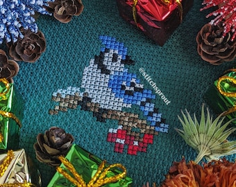 May include: A bluebird cross-stitch pattern on a green fabric background. The design includes a bluebird with a red breast, surrounded by pine cones, pine branches, and small wrapped gifts.