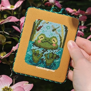 May include: A cross-stitch embroidery of two green frogs sitting in a pond with lily pads. The embroidery is framed in a yellow and green border with the text "@stitchsprout" in the bottom right corner.