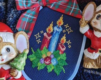 May include: A blue and white cross-stitch ornament featuring a candle and wreath design. The ornament is surrounded by two ceramic mice wearing Santa hats. The ornament is decorated with a red and green tartan bow.