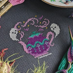 以下が含まれることがあります： 紫色のティーポットに緑色の蓋と白い幽霊がついたクロスステッチパターン。ティーポットは星に囲まれ、その上に「Boo」という文字が刺繍されています。クロスステッチは濃いグレーの生地に施されています。