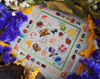 May include: A cross-stitch pattern featuring a colorful array of mushrooms, ladybugs, and other woodland creatures. The design is framed by a border of green and yellow leaves. The cross-stitch is finished and displayed on a white background.