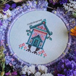 May include: A cross-stitch embroidery of a whimsical house with a blue and green exterior, pink and yellow accents, and a small door. The house is on stilts with flowers around it. The embroidery is framed in a purple and white crocheted hoop with the text "@stitchsprout" in the upper right corner.