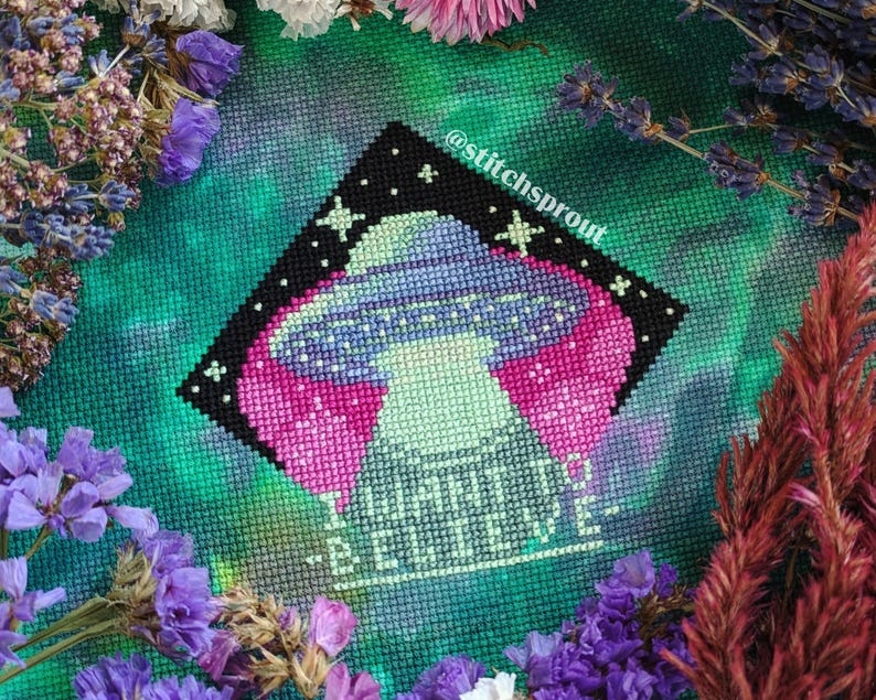 K&ouml;nnte beinhalten: Kreuzstichmuster mit einem UFO mit einem rosa und violetten Design und dem Text "Glaub nicht" in einer gr&uuml;nen und wei&szlig;en Schrift. Das Muster ist von Blumen und Gr&uuml;n umgeben.