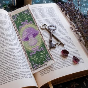 May include: A cross-stitch bookmark featuring a purple mushroom with green leaves and a white background. The bookmark has a fringed edge and the text "@stitchsprout" is visible in the lower right corner.
