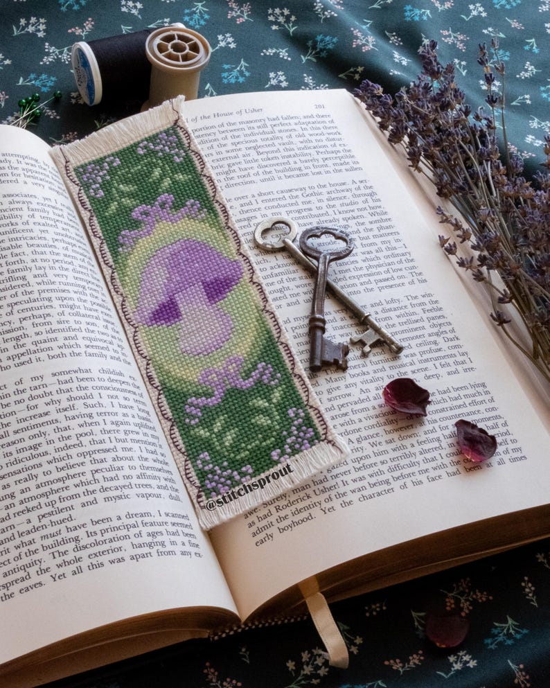 May include: A cross-stitch bookmark featuring a purple mushroom with green leaves and a white background. The bookmark has a fringed edge and the text "@stitchsprout" is visible in the lower right corner.