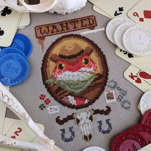 May include: Cross-stitch pattern of a fox wearing a cowboy hat and bandana, with the words "WANTED" and "UTU" stitched above and below the fox. The pattern is surrounded by playing cards and poker chips.