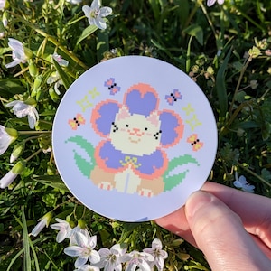 May include: A sticker featuring a pixelated cat wearing a flower costume. The cat is white with pink cheeks and a purple flower with pink petals. The background is light blue with yellow and green accents.