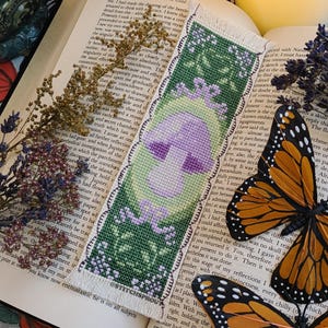 May include: A cross-stitch bookmark featuring a purple mushroom with a green stem and leaves. The bookmark has a white border with a scalloped edge and the text "ESTITCHSPROUT" at the bottom.