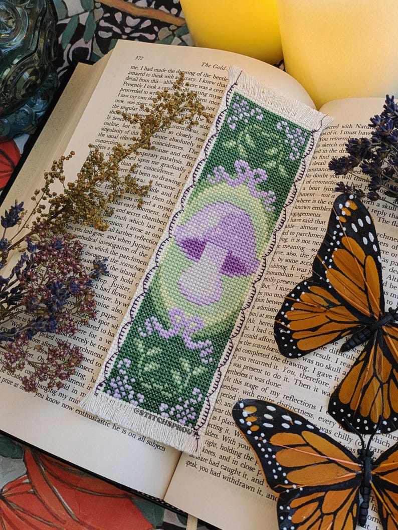 May include: A cross-stitch bookmark featuring a purple mushroom with a green stem and leaves. The bookmark has a white border with a scalloped edge and the text "ESTITCHSPROUT" at the bottom.