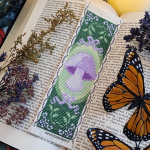 May include: A purple and green cross-stitch bookmark featuring a mushroom with a white stem and a green background. The bookmark has a scalloped edge and the text "ESTITCHSPROUT" is printed at the bottom.