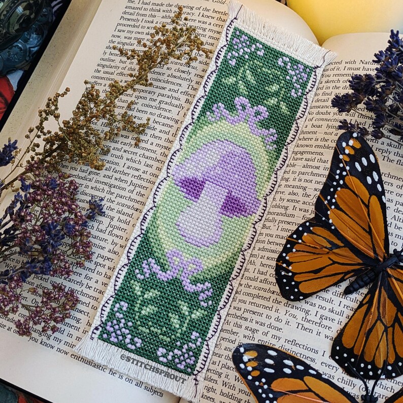 May include: A purple and green cross-stitch bookmark featuring a mushroom with a white stem and a green background. The bookmark has a scalloped edge and the text "ESTITCHSPROUT" is printed at the bottom.