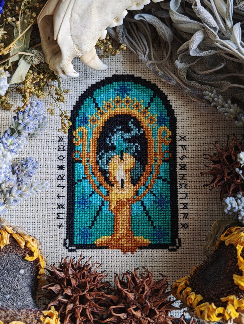 May include: A cross-stitch embroidery of a stylized tree with a golden trunk and blue and green leaves. The tree is framed in a black arch with a gold border. The background is a dark blue color. The embroidery is surrounded by dried flowers and other natural elements.