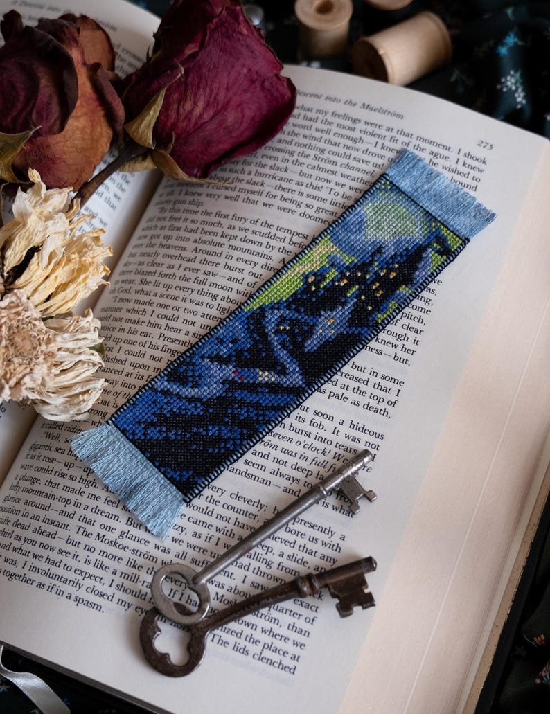 May include: A handmade bookmark with a cross-stitch design depicting a dark landscape with a starry sky. The bookmark has a blue fringe and is placed on an open book with dried roses and antique keys.