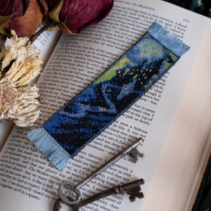 May include: A handmade bookmark with a cross-stitch design depicting a dark, mountainous landscape under a starry sky. The bookmark has a light blue fringe and is placed on an open book.