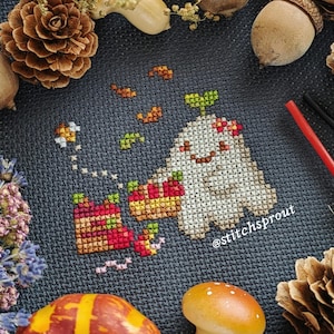 May include: Cross-stitch pattern of a ghost with a smiling face, wearing a crown of leaves and holding a basket of apples. The pattern is surrounded by autumn leaves and acorns. The text "@stitchsprout" is in the bottom right corner.