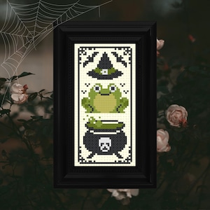 Puede incluir: Un diseño de punto de cruz enmarcado que presenta una rana verde, un caldero con poción verde y un sombrero de bruja con un borde verde. El diseño está sobre un fondo blanco con acentos negros.