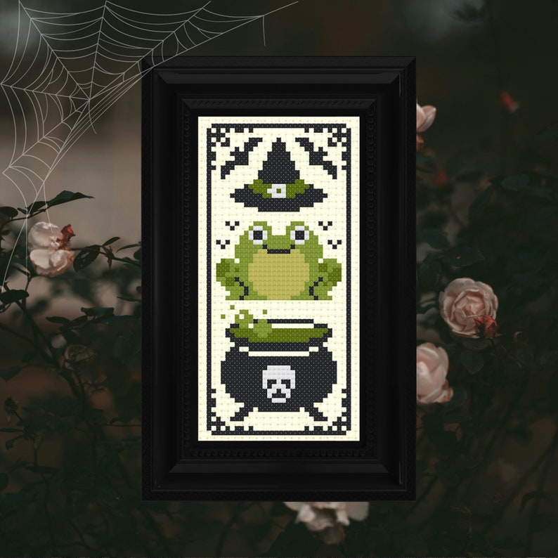 K&ouml;nnte beinhalten: Ein gerahmtes Kreuzstich-Design mit einem gr&uuml;nen Frosch, einem Kessel mit gr&uuml;nem Zaubertrank und einem Hexenhut mit gr&uuml;nem Rand. Das Design ist auf wei&szlig;em Hintergrund mit schwarzen Akzenten.