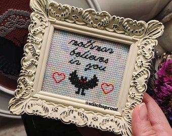 May include: A white framed cross-stitch with a black bat and the text "no one believes in you" in black thread. Two red hearts are stitched on either side of the bat. The cross-stitch is on a light blue background with a rainbow pattern. The frame is ornate and has a distressed white finish. The username "@stitchsprout" is stitched in black thread at the bottom of the cross-stitch.