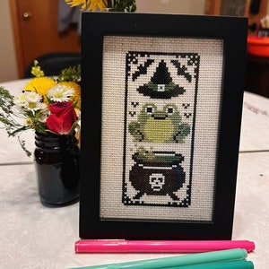 K&ouml;nnte beinhalten: Ein gerahmtes Kreuzstich-Design mit einem gr&uuml;nen Frosch, der einen Hexenhut tr&auml;gt und auf einem Kessel mit Totenkopf und Knochen sitzt. Der Text "FINISH @SUSIE_BRUISER PATTERN @STITCHSPROUT" befindet sich unter dem Bild.