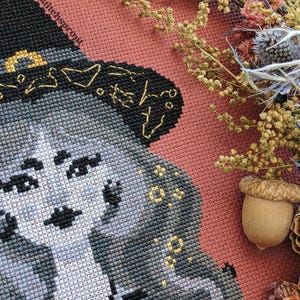 Op de afbeelding: Een kruissteekportret van een vrouw met een heksenhoed, met grijs haar en een zwarte hoed. De kruissteek staat op een roze stoffen achtergrond, met gedroogde bloemen en een eikel in de hoek. De tekst "@stitchesprout" is zichtbaar.