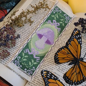 May include: A green and purple cross-stitch bookmark with a mushroom design and the text "ESTITCHSPROUT". Two orange and black monarch butterflies are on a book page with text.