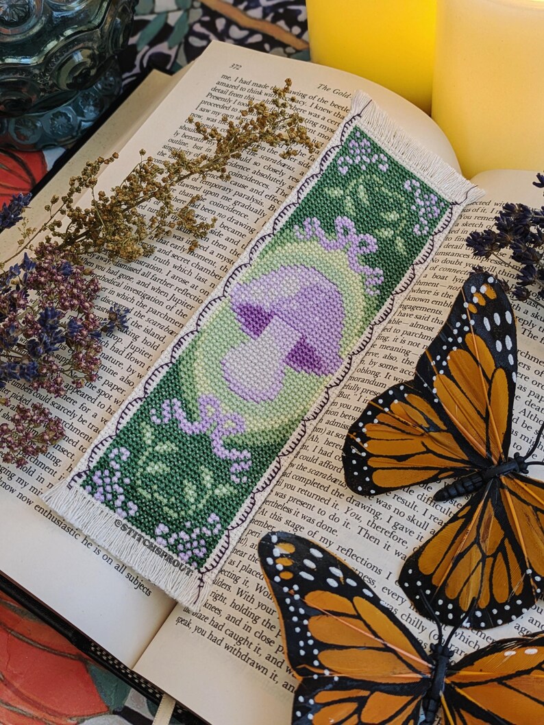 May include: A green and purple cross-stitch bookmark with a mushroom design and the text "ESTITCHSPROUT". Two orange and black monarch butterflies are on a book page with text.