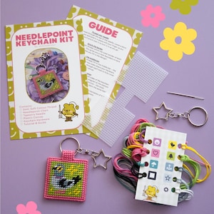 May include: A needlepoint keychain kit featuring a printed guide, a clear plastic canvas, a metal tapestry needle, a star-shaped keychain ring, and a thread organizer with various colored cotton threads. A finished square keychain with a bird design is included.