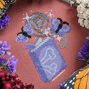 Puede incluir: Bordado de punto de cruz morado y gris de una botella con un diseño de mariposa. El bordado está sobre un fondo rojo.