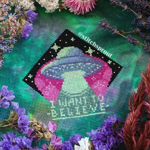 K&ouml;nnte beinhalten: Kreuzstichmuster eines gr&uuml;nen Au&szlig;erirdischen mit einem rosa und violetten Raumschiff, umgeben von Sternen. Der Text "What's up, Earth?" ist unter dem Au&szlig;erirdischen gestickt. Das Muster ist von einem gr&uuml;nen und blauen Hintergrund mit einem wei&szlig;en Rand umgeben.