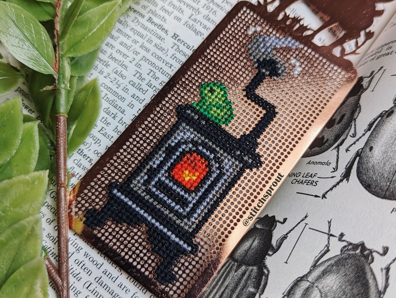 K&ouml;nnte beinhalten: Ein kupferfarbenes Lesezeichen mit einem schwarzen und wei&szlig;en Kreuzstich-Design eines kleinen Ofens mit Rauch aus dem Schornstein. Ein gr&uuml;ner Frosch sitzt auf dem Ofen. Das Lesezeichen hat den Text "@stitchsprout" aufgedruckt.