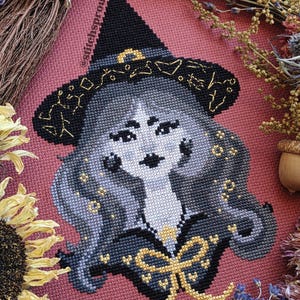 Op de afbeelding: Kruissteekontwerp van een heks met een zwarte hoed met gouden accenten en een zwart-gouden outfit. De heks heeft grijs haar en een strenge uitdrukking. De achtergrond is een roze stof. De tekst "stitchsprout" is zichtbaar.