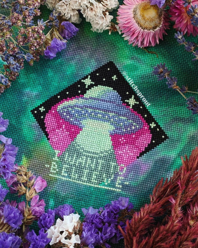 K&ouml;nnte beinhalten: Kreuzstichmuster eines UFOs mit Sternen und dem Text "I have to believe" auf einem gr&uuml;n-violetten Tie-Dye-Hintergrund.