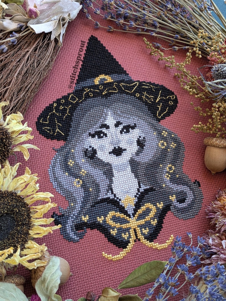 Op de afbeelding: Kruissteekportret van een heks met een zwarte hoed met gouden details. De heks heeft grijs haar en een zwarte outfit met een gouden strik. De achtergrond is roze stof, omlijst door gedroogde bloemen en zonnebloemen.