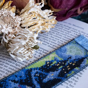 May include: A handmade bookmark with a cross-stitch design featuring a night sky scene in shades of blue, green, and black. The bookmark is placed on an open book, with dried flowers in the background.