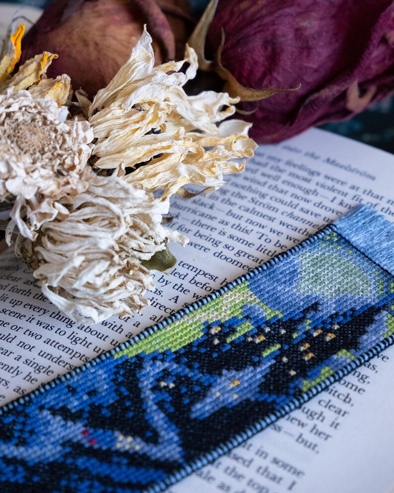 May include: A handmade bookmark with a cross-stitch design featuring a night sky scene in shades of blue, green, and black. The bookmark is placed on an open book, with dried flowers in the background.