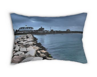 May include: Rectangular pillow featuring a coastal scene. A stone pier extends into the water, leading to buildings on the shore under a cloudy sky. The pillow has a blue and gray color palette, evoking a serene seaside atmosphere.