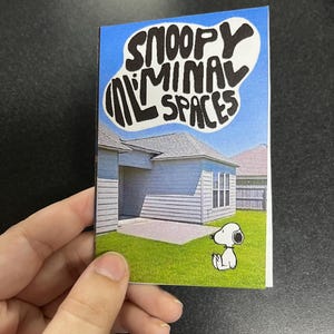 May include: A small card featuring a cartoon illustration of Snoopy from Peanuts. The card has the text "Snoopy Minimal Spaces" in a stylized font. The background shows a house and a blue sky.