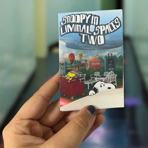 May include: A small rectangular card with the text "Snoopy in Liminal Spaces Two" and an illustration of Snoopy and Woodstock in a park with hot air balloons. The card is held in a person's hand.