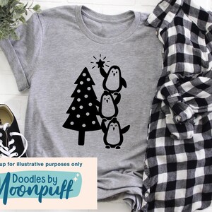 K&ouml;nnte beinhalten: Ein meliertes graues T-Shirt mit einer schwarzen Grafik eines Weihnachtsbaums und drei Pinguinen. Ein Pinguin h&auml;lt einen Stern. Das Shirt hat auch den Text "Doodles by Moonpuff" am unteren Rand.