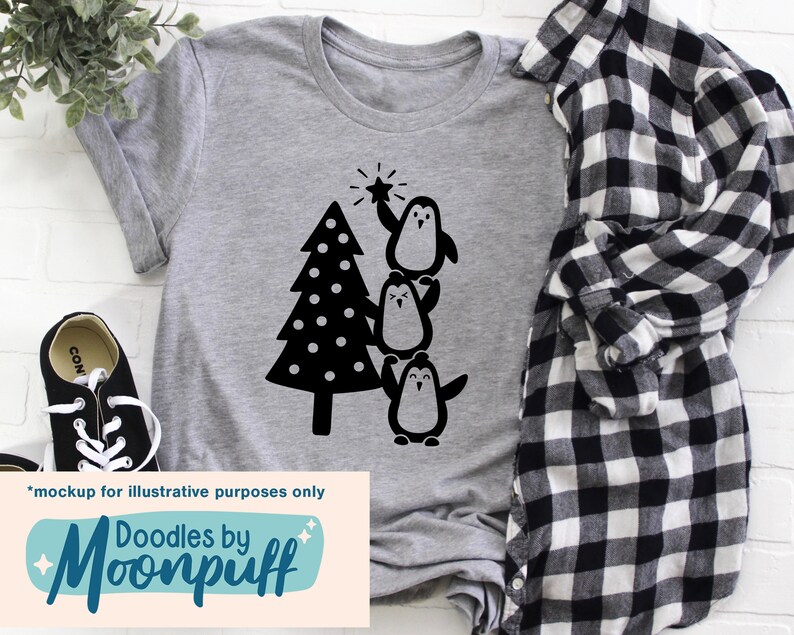 K&ouml;nnte beinhalten: Ein meliertes graues T-Shirt mit einer schwarzen Grafik eines Weihnachtsbaums und drei Pinguinen. Ein Pinguin h&auml;lt einen Stern. Das Shirt hat auch den Text "Doodles by Moonpuff" am unteren Rand.
