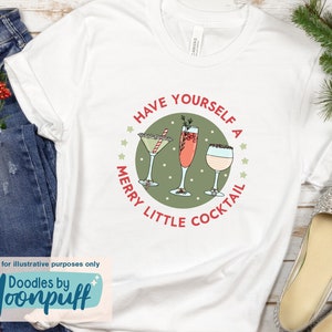 May include: White t-shirt with a festive cocktail design. The design features three cocktail glasses inside a green circle with the text "Have Yourself a Merry Little Cocktail". The shirt has a rolled sleeve and is displayed with holiday decor.