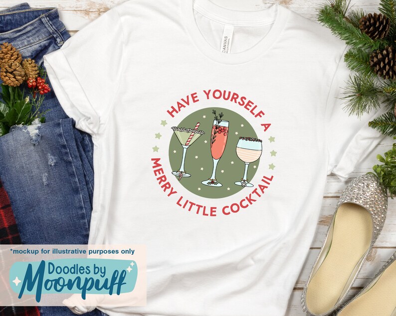 May include: White t-shirt with a festive cocktail design. The design features three cocktail glasses inside a green circle with the text "Have Yourself a Merry Little Cocktail". The shirt has a rolled sleeve and is displayed with holiday decor.